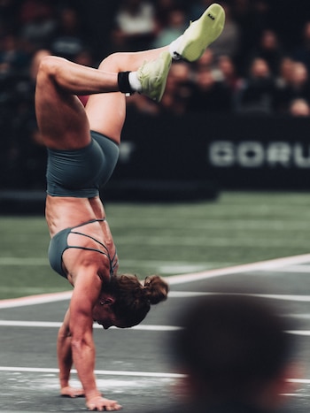 Una mujer con ropa deportiva gris camina sobre las manos con el cuerpo invertido y las piernas en el aire en una pista cubierta
