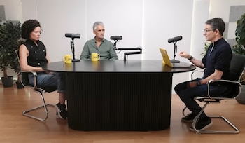 Tres personas sentadas alrededor de una mesa ovalada negra con tres micrófonos, dos tazas amarillas y una computadora portátil amarilla. Hay plantas