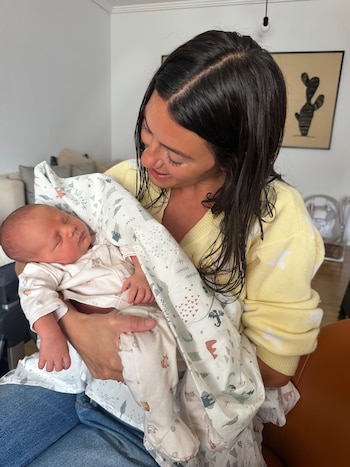 Sofía Jujuy Jiménez sostiene a un bebé recién nacido dormido, envuelto en una manta blanca con estampados. Viste un suéter amarillo y jeans azules