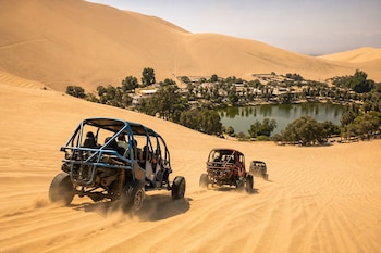 Tres vehículos tubulares con pasajeros descienden por una duna de arena frente al oasis de Huacachina y su laguna.