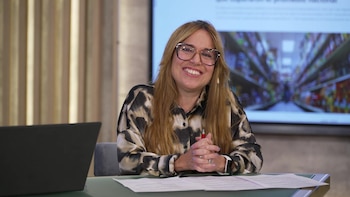 Mujer rubia con gafas y camisa estampada sentada en un escritorio frente a un portátil. Detrás, una gran pantalla muestra un pasillo de supermercado con estantes llenos