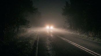 Carretera nocturna de asfalto rodeada de árboles con una densa capa de niebla y dos faros encendidos en el fondo.