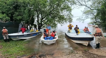 Tres lanchas pequeñas con personas que llevan chalecos salvavidas en una orilla arbolada con el suelo anegado