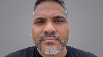 Primer plano del rostro de un hombre con barba canosa y camiseta negra frente a un fondo gris.