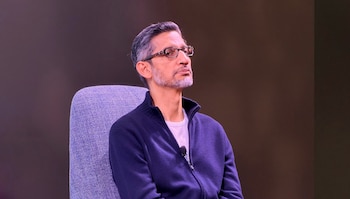 El CEO de Google, Sundar Pichai, sentado en un sillón gris, con gafas, barba y cabello entrecano, vistiendo una chaqueta azul marino con cremallera sobre una camiseta blanca