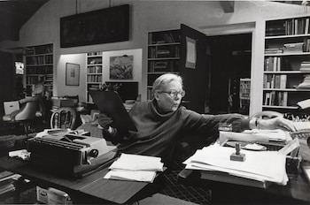 Fotografía en blanco y negro de una mujer con gafas sentada en un escritorio con una máquina de escribir, documentos, y estanterías llenas de libros