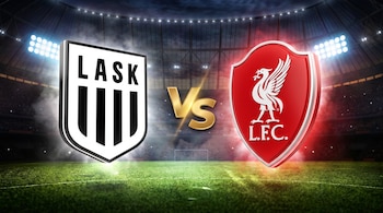Un escudo blanco y negro del LASK y un escudo rojo del Liverpool flanquean las letras doradas VS sobre el campo de un estadio iluminado.