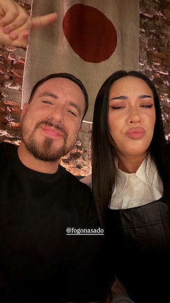 Fede Bal sonríe mientras Evelyn Botto posa con labios fruncidos en una selfie. Detrás, una pared de ladrillos y un tapiz beige con círculo rojo