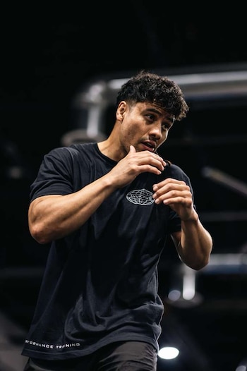 Hombre de cabello oscuro y camiseta negra con logotipo Invictus Performance Training, pantalón oscuro, con manos cerca del rostro, fondo desenfocado