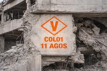 Estructura de hormigón gris con daños severos, varillas de metal expuestas. Señal naranja pintada en la pared: un rombo con una V, COL01 y 11 AGOS.