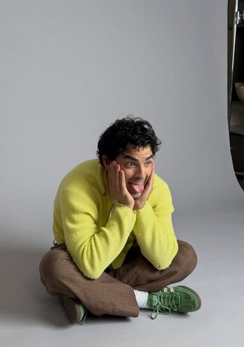 Joe Jonas sentado en el suelo con cárdigan amarillo y pantalones marrones, sacando la lengua con las manos en la cara, sobre un fondo gris claro