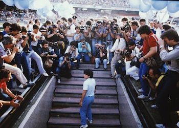 Diego Maradona de espaldas sube escaleras en un estadio. Numerosos fotógrafos con cámaras, flashes y lentes lo rodean. Multitud en las gradas y globos azules