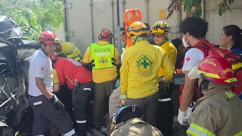 Varios socorristas con uniformes blancos y rojos inspeccionan un automóvil negro destruido junto a la estructura de un camión