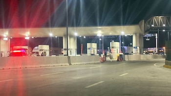 Una patrulla de la Guardia Nacional con luces rojas intermitentes en una caseta de peaje iluminada de una carretera por la noche, con camiones y figuras humanas