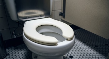 Vista frontal de un inodoro de porcelana blanca con asiento de apertura frontal, ubicado en un baño con piso de baldosas hexagonales grises y paredes claras.