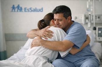 Un hombre abraza a una mujer en una habitación de hospital. Ella viste una bata blanca, él un uniforme azul. Se observa un logo de EsSalud y equipo médico.