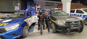 Hombre esposado con dos agentes policiales, dos camionetas policiales azules y negras, un SUV policial blanco y un camión de carga