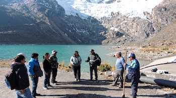 Ocho personas de pie conversan cerca de una laguna turquesa con un glaciar y montañas nevadas en el fondo.