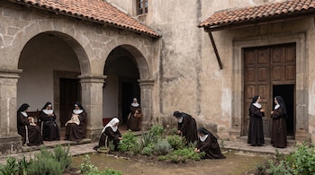 Varias monjas con hábitos negros y blancos en un patio de convento de piedra con arcos. Algunas bordan, otras cultivan plantas y dos conversan.