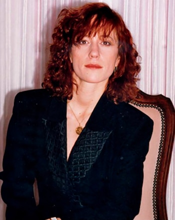 Mujer de cabello rojizo y rizado, con blazer oscuro y collar dorado, sentada frente a una cortina de rayas verticales claras, mirando al frente