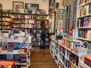 Interior de librería con estanterías de madera y blancas repletas de libros, una persona subida a una escalera, varias ventanas y cuadros decorativos