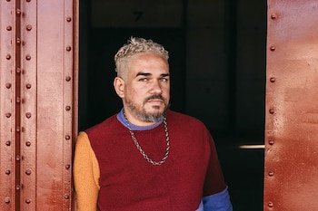 Retrato de Raphael Fonseca, hombre con cabello rizado claro y barba, usando suéter multicolor y cadena, apoyado en una pared metálica rojiza