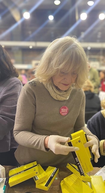 Mujer de cabello rubio con guantes blancos organiza cajas amarillas de Losacor A 100. Lleva un suéter beige y un pin rosa de 'Navarro Viola Voluntaria'