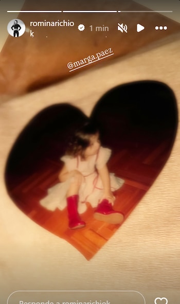 Una niña sentada en un suelo de madera, vestida con un vestido blanco y botas rojas, vista a través de un desenfoque en forma de corazón
