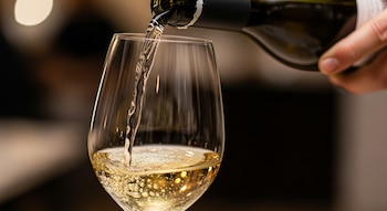Una mano sostiene una botella de vino negro y vierte vino blanco dorado en una copa de cristal transparente, con burbujas visibles.