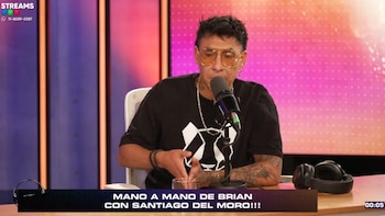 Brian Sarmiento, con gafas de sol y camiseta negra, sentado frente a un micrófono en un estudio de streaming con luces de colores y auriculares sobre la mesa