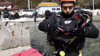 Un hombre con traje de buceo negro y gorro naranja revisa sus accesorios de inmersión a la orilla del agua