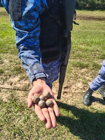 Primer plano de la mano de un agente de policía con uniforme camuflado azul, sosteniendo cinco proyectiles esféricos de tierra. Un arma y otra pierna policial visibles