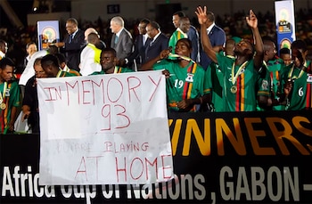Campo de fútbol con varios jugadores de Zambia en uniforme verde, portando medallas y sosteniendo una pancarta blanca con el texto 'IN MEMORY of 93'