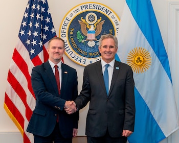 Dos hombres en traje se dan la mano ante el sello de la Oficina del Representante Comercial y las banderas de Estados Unidos y Argentina