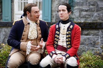 Brandon Scott Jones y John Hartman, vestidos con trajes militares coloniales, sentados al aire libre frente a un muro de piedra y vegetación
