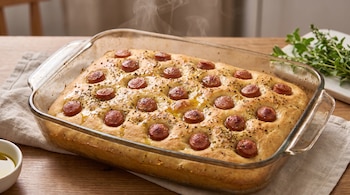 Focaccia horneada con rodajas de salchicha en una fuente de vidrio, un recipiente con aceite de oliva y ramas de orégano sobre un mantel.