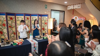 Un hombre con chaqueta de chef y una mujer con delantal azul están detrás de una mesa con ollas y comida. Un público aplaude frente a ellos. Fondo de pantallas florales