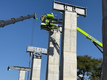 Imagen de un trabajador en una plataforma elevadora junto a grandes pilares de concreto y estructuras metálicas grises en construcción, con una grúa y cielo azul