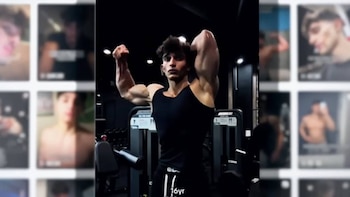 Un hombre joven de piel clara con camiseta sin mangas negra, Performance M, flexiona sus bíceps y tríceps en un gimnasio oscuro con máquinas de ejercicio