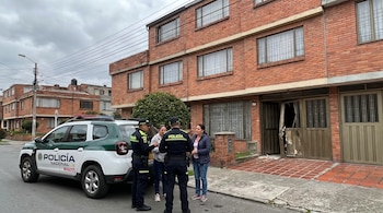 Dos oficiales de la Policía Nacional junto a su vehículo conversan con dos mujeres frente a una casa de ladrillo rojo con una puerta dañada en Bogotá.
