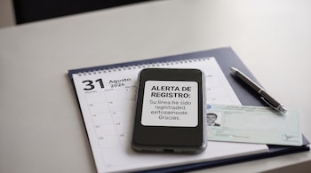 Un teléfono celular moderno enciende su pantalla con una alerta de registro de línea, sobre un calendario, una identificación oficial y un bolígrafo en una mesa.