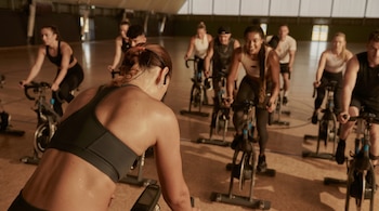 Vista trasera de una mujer sudando en una clase de spinning; otras personas en bicicletas estáticas se ven en el fondo de un gimnasio.