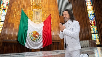 Hombre con barba arrodillado, manos juntas, traje blanco con brillos, frente a una bandera de México y un cuadro de la Virgen de Guadalupe en una iglesia con vitrales.