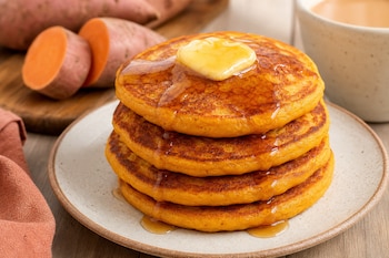 Cuatro hot cakes apilados en un plato con mantequilla y jarabe, junto a camotes cortados sobre una tabla de madera.