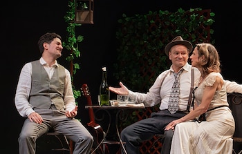 Tres actores sentados en un escenario teatral. Un hombre de chaleco, un hombre de sombrero y corbata, y una mujer de vestido. Una mesa con botella, vasos y una guitarra. Fondo con celosía, hiedra y lámpara