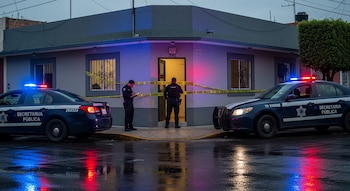 Patrullas de policía con luces encendidas y dos oficiales inspeccionan una casa acordonada con cinta amarilla en una calle mojada al anochecer.