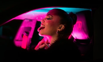 Wanna, de perfil, sacando la lengua, con maquillaje brillante y un collar, en un coche oscuro. Iluminada por luces de neón rosas y azules