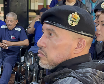 Un hombre de perfil viste boina negra con el escudo de Venezuela, mientras al fondo un señor permanece en silla de ruedas
