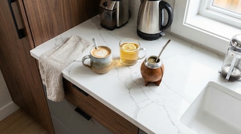 Una vista superior de una encimera de cuarzo blanco en una cocina moderna, mostrando una taza de café, un vaso de té con limón y un mate de calabaza con bombilla.