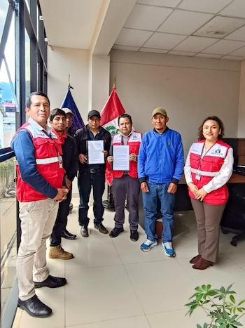 Un grupo de siete personas, incluyendo a cuatro hombres y tres con chalecos rojos de Cofopri, posan en una oficina con banderas peruanas. Dos hombres sostienen documentos